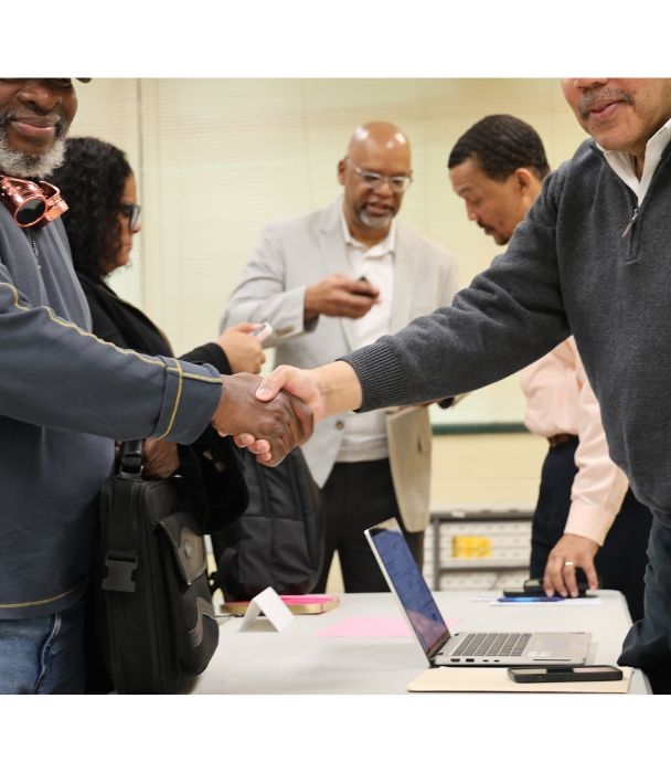 Two men shaking hands over a table with others in the background talking together.