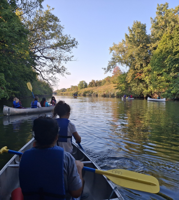 People in canoes paddling down a calm river surrounded by lush green trees.