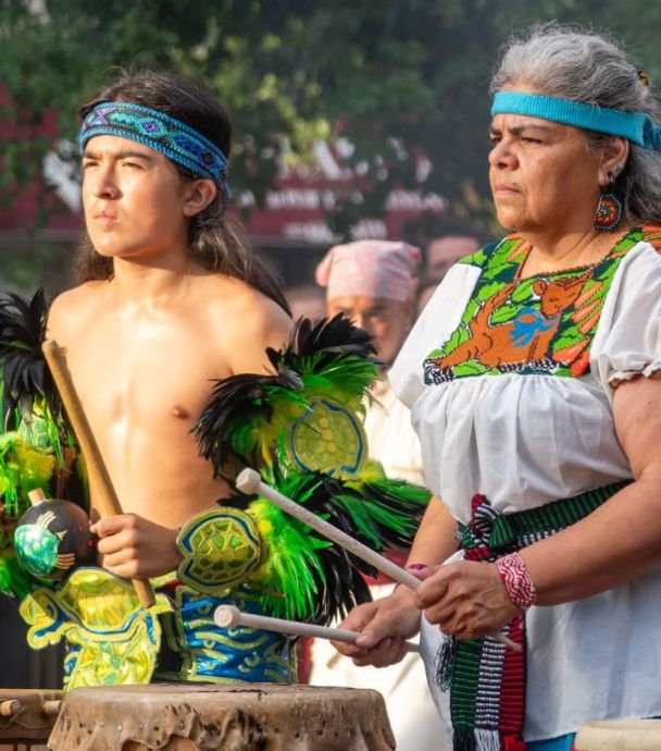 Photograph of two indigenous people playing drums in traditional ceremonial clothing.